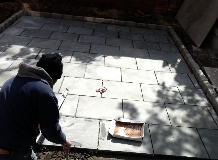 Concrete Pavers Brooklyn team is working on a residential house. In this image you see a Brooklyn concrete paver standing on his knees with a hammer in his hand. This image shows an outside patio with approximately 400 sq ft. This project took about 2 days to finish. The image was taken in April 2022.
